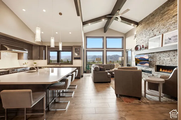 Open-concept living area featuring a vaulted ceiling with exposed beams, expansive window array, wood-finish flooring, a stone-faced fireplace, and a kitchen island with seating