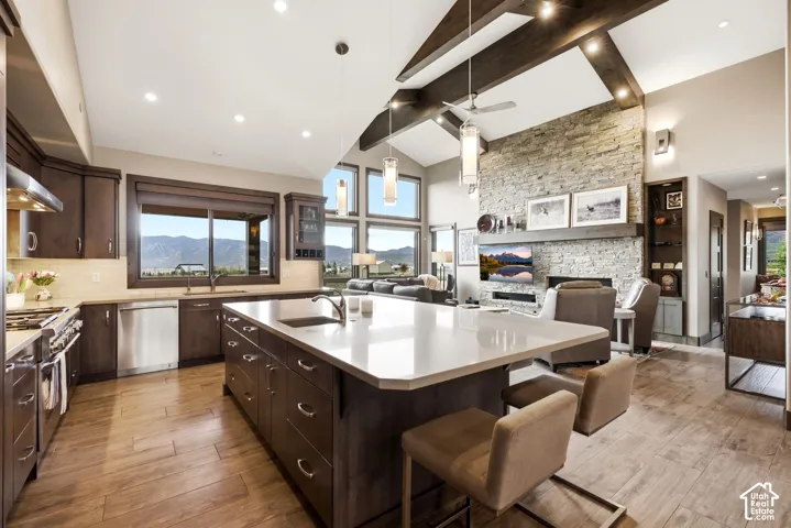 Open-concept kitchen and living area featuring a stone-clad fireplace, vaulted ceiling with exposed beams, wood-finish flooring, and a kitchen island with seating