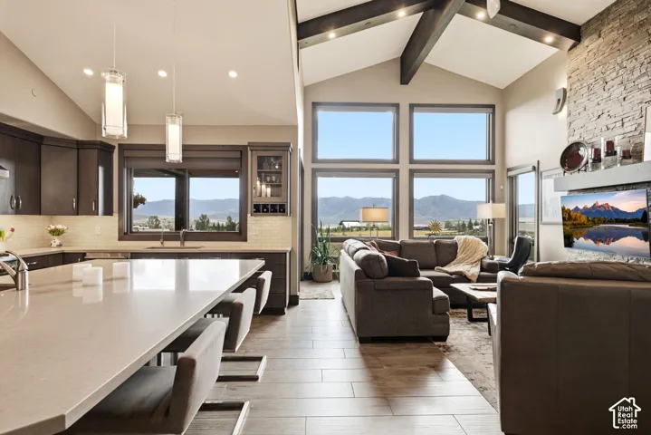 Vaulted ceiling with exposed beams, stone-clad fireplace, large picture windows, wood-finish flooring, and a spacious kitchen island with seating