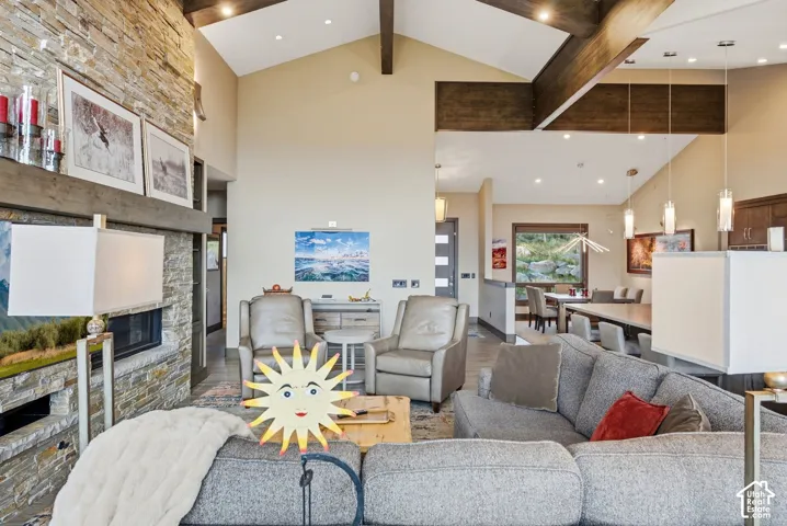 Vaulted ceilings with exposed wood beams, a stone-clad fireplace with a dark wood mantel, recessed lighting, and a dining area with a window