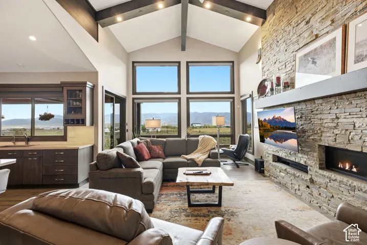 Vaulted ceiling with exposed wood beams, expansive windows with mountain scenery, stone-clad fireplace, built-in cabinetry, and wood-finish flooring