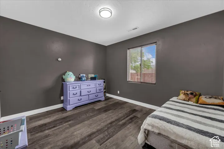 Spacious bedroom featuring warm gray walls, wood-finish flooring, a large window with plantation shutters, and a purple six-drawer dresser with decorative hardware