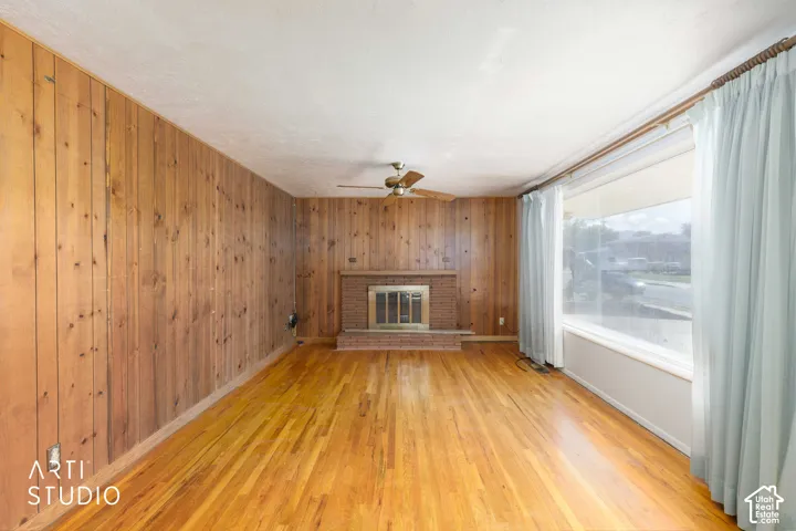 Wood-paneled wall, brick fireplace with metal insert, wood-finish flooring, ceiling fan, and large picture window