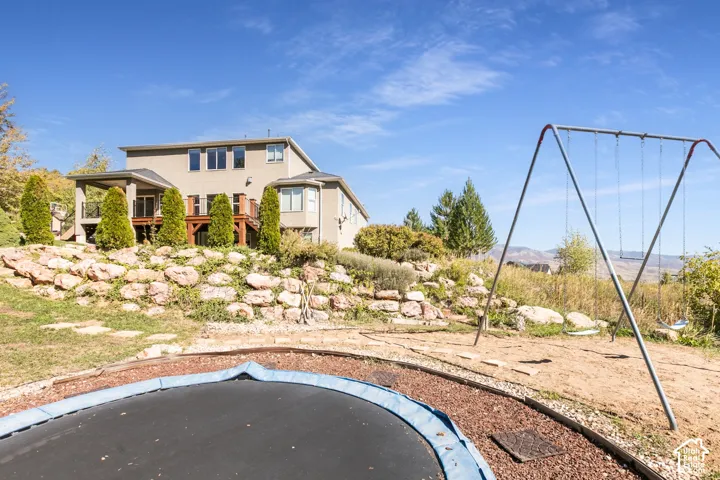 Spacious exterior featuring a tan stucco finish, multiple windows, a covered patio, a wood deck, and a terraced rock garden with established plantings