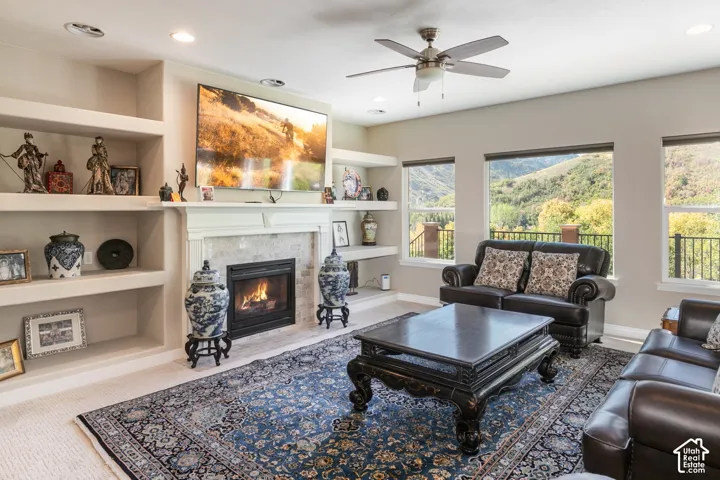Built-in shelving, stone fireplace with a white mantel, recessed lighting, ceiling fan, and multiple windows with a natural landscape