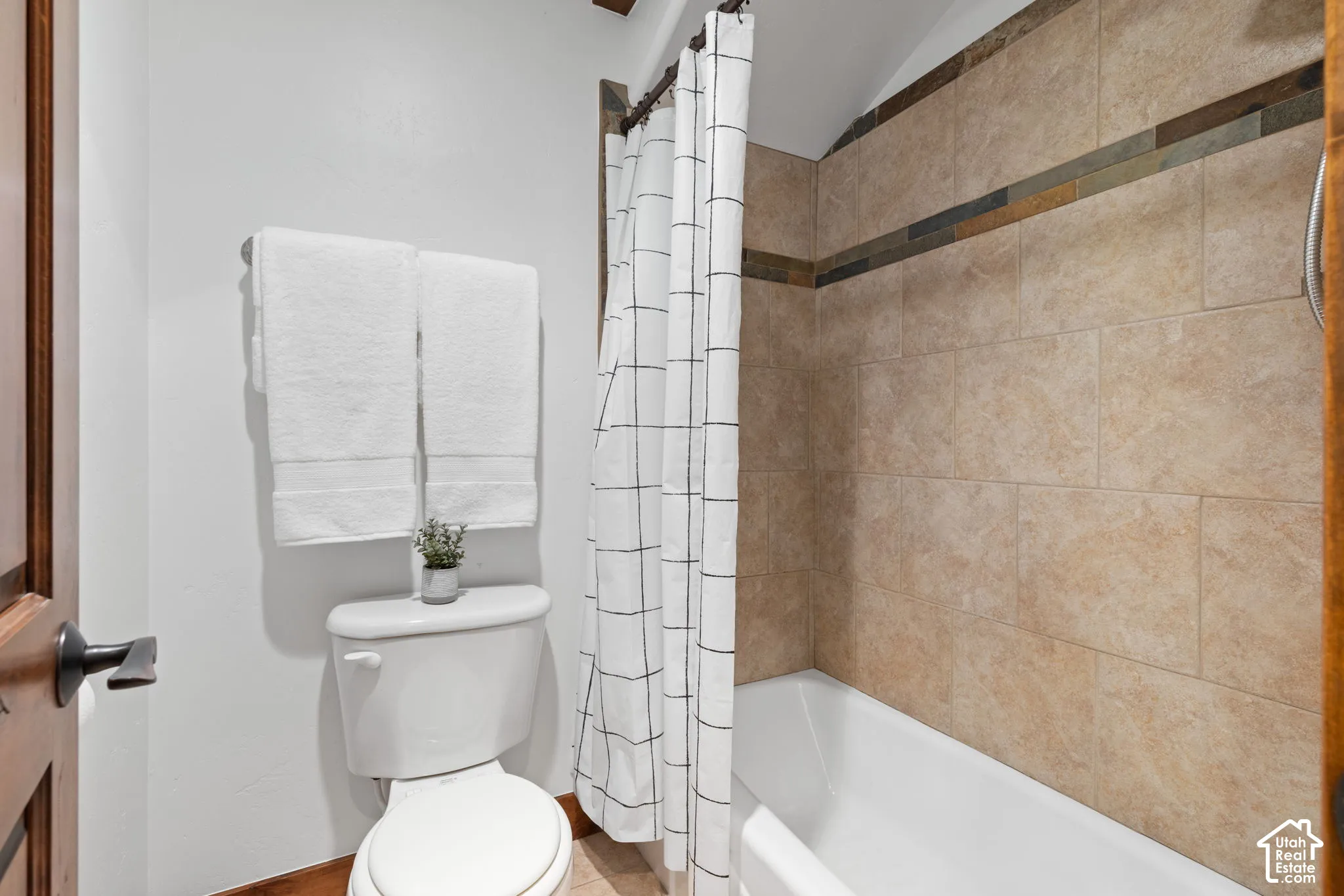 Tiled shower surround with decorative accent band, white bathtub, and wood-finish door with dark hardware