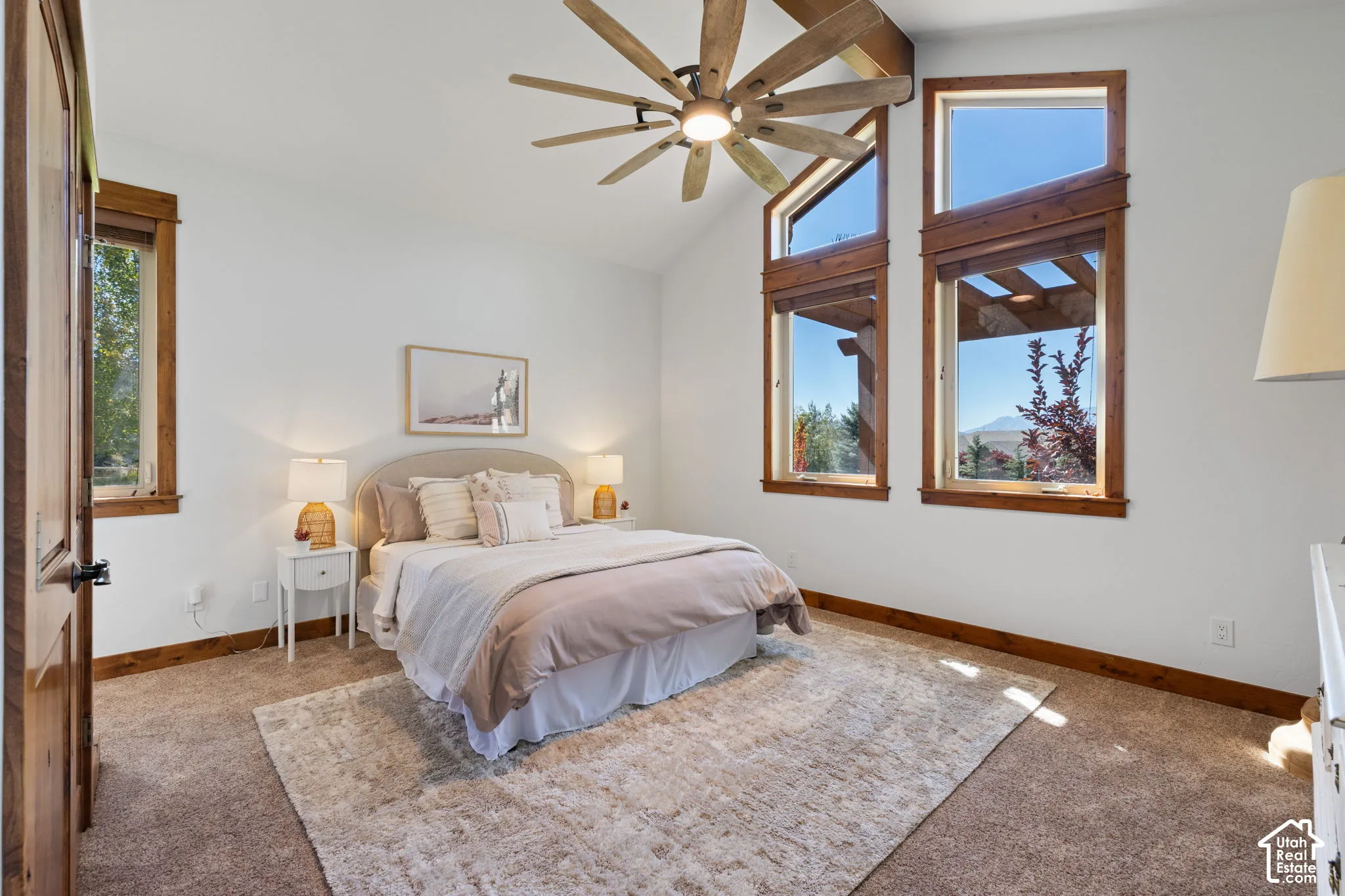 Vaulted ceilings with exposed wood beams, wood-trimmed windows, a multi-blade ceiling fan, and light-toned carpet flooring