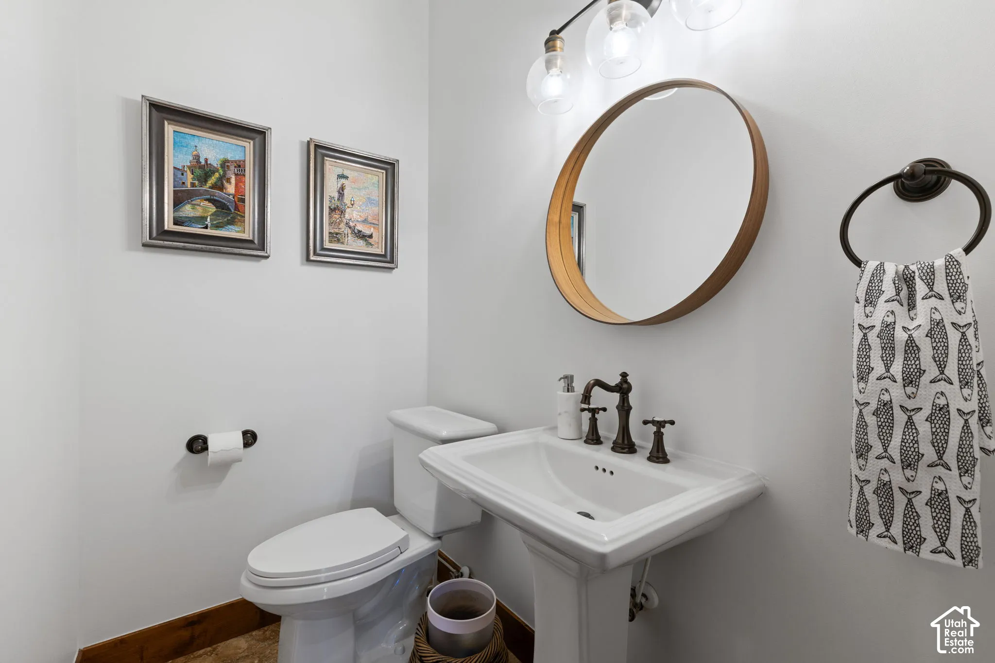 Pedestal sink with dark bronze fixtures, circular mirror with wood-finish frame, and overhead vanity lighting