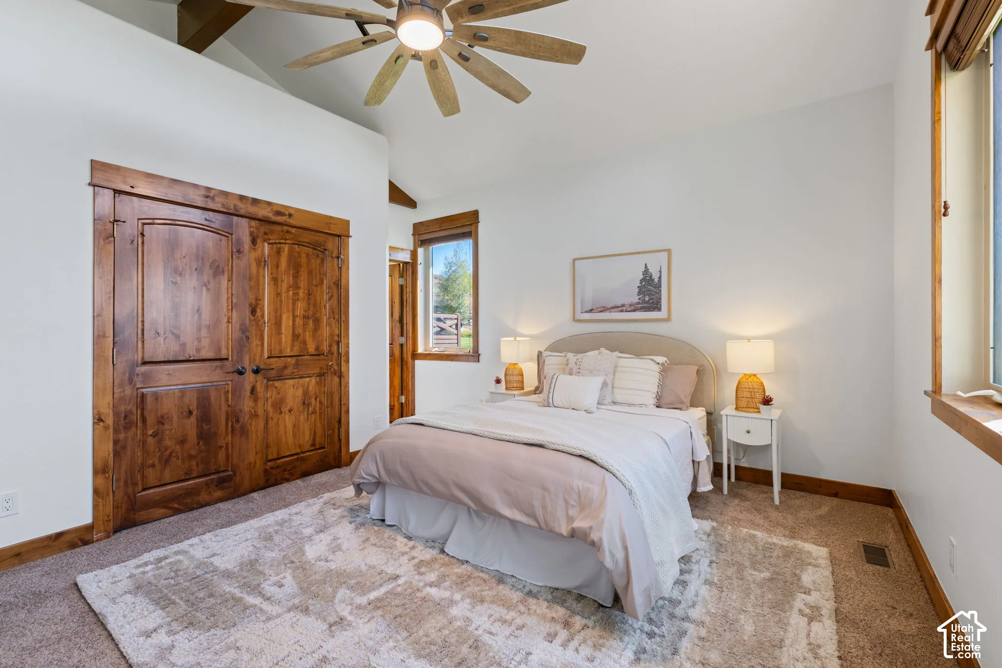 Vaulted ceiling with exposed wood beams, wood-finish ceiling fan, wood-paneled closet doors, carpeted flooring, and a large window with wood trim