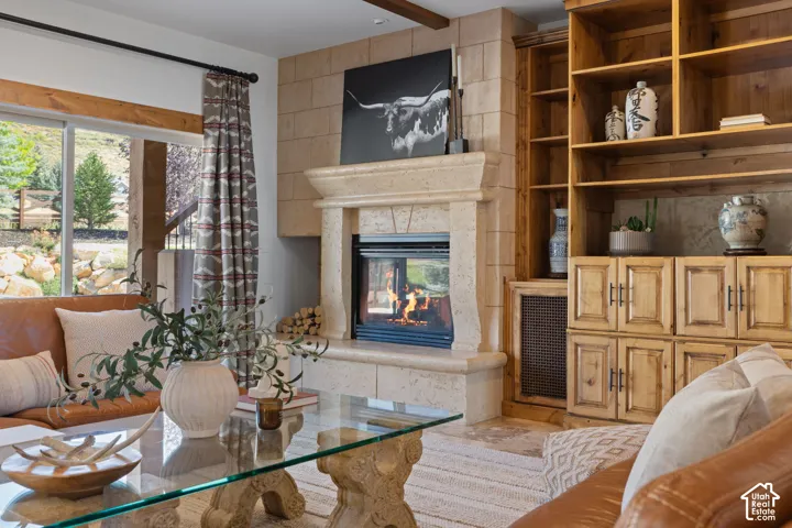 Stone-clad fireplace with hearth, integrated wooden built-in shelving, glass-top coffee table with stone base, and a large window providing ample natural illumination