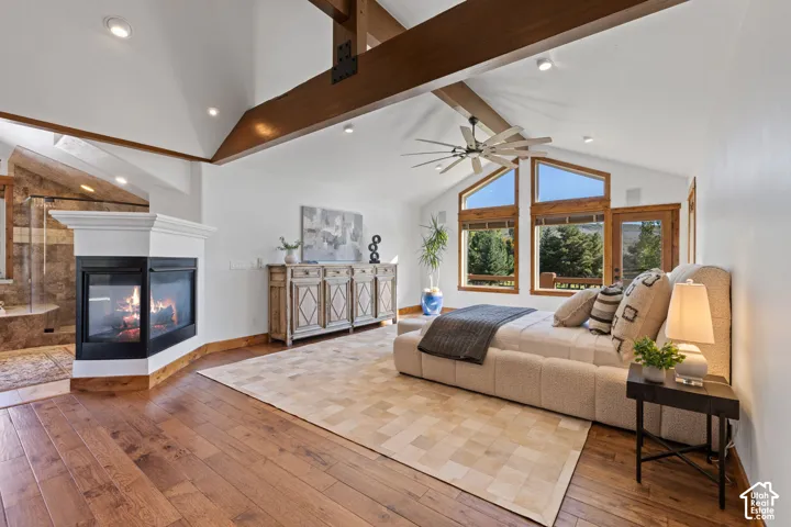 Vaulted ceilings featuring exposed wood beams, a corner fireplace with a white mantel, expansive wood-framed windows, wood-finish flooring, and a ceiling fan