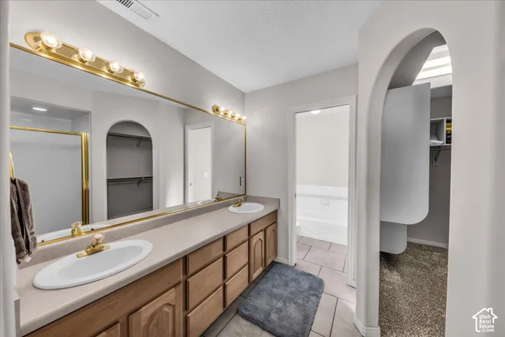 Dual vanity with gold-tone hardware, full-width mirror, wood-finish cabinetry, and an arched entryway to a walk-in closet