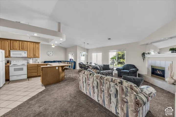 Open-concept living and kitchen area featuring ceramic tile flooring, wood-finish cabinetry with white appliances, a center island with seating, recessed lighting, a fireplace with surround shelving, and abundant natural light from multiple windows