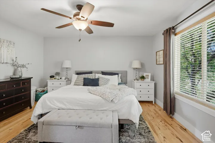 Hardwood flooring, ceiling fan with integrated lighting, window with blinds, light gray wall finishes, and a neutral upholstered headboard