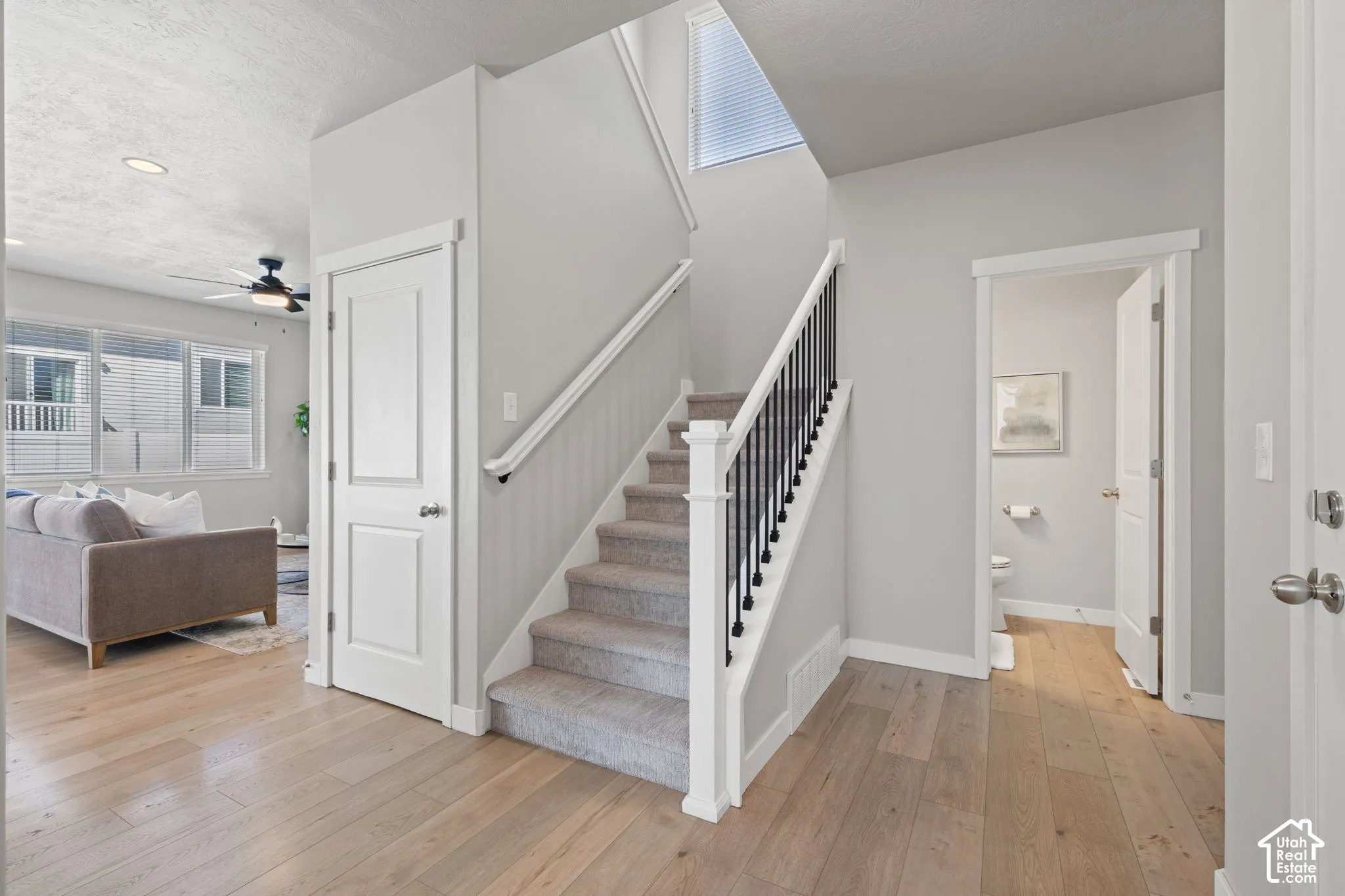 Wood-finish flooring, staircase with carpeted treads and black metal balusters, and a ceiling fan with integrated lighting