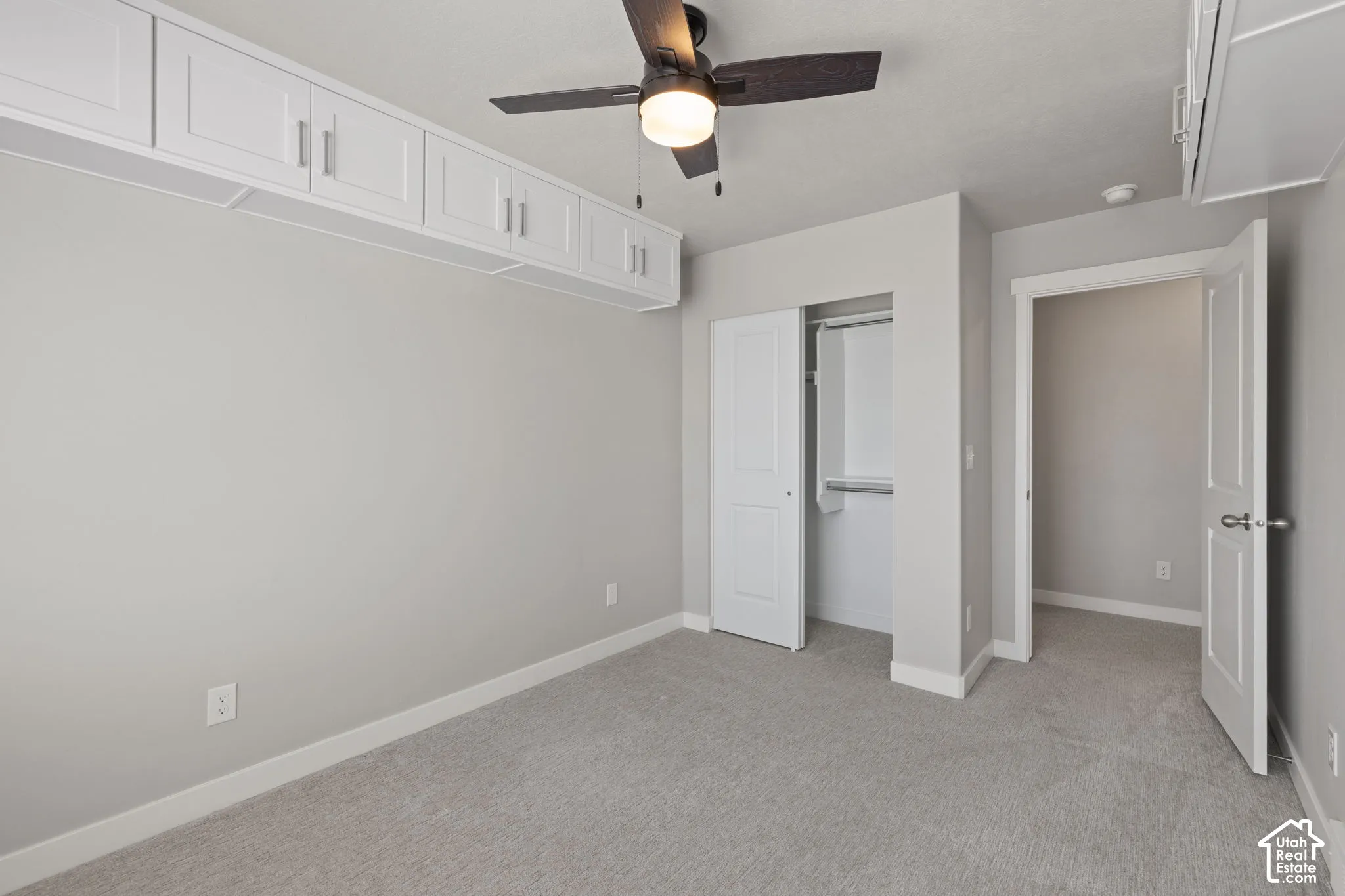 Recessed overhead cabinetry with brushed nickel hardware, ceiling fan with integrated lighting, soft grey wall paint, plush grey carpeting, and a bi-fold door closet