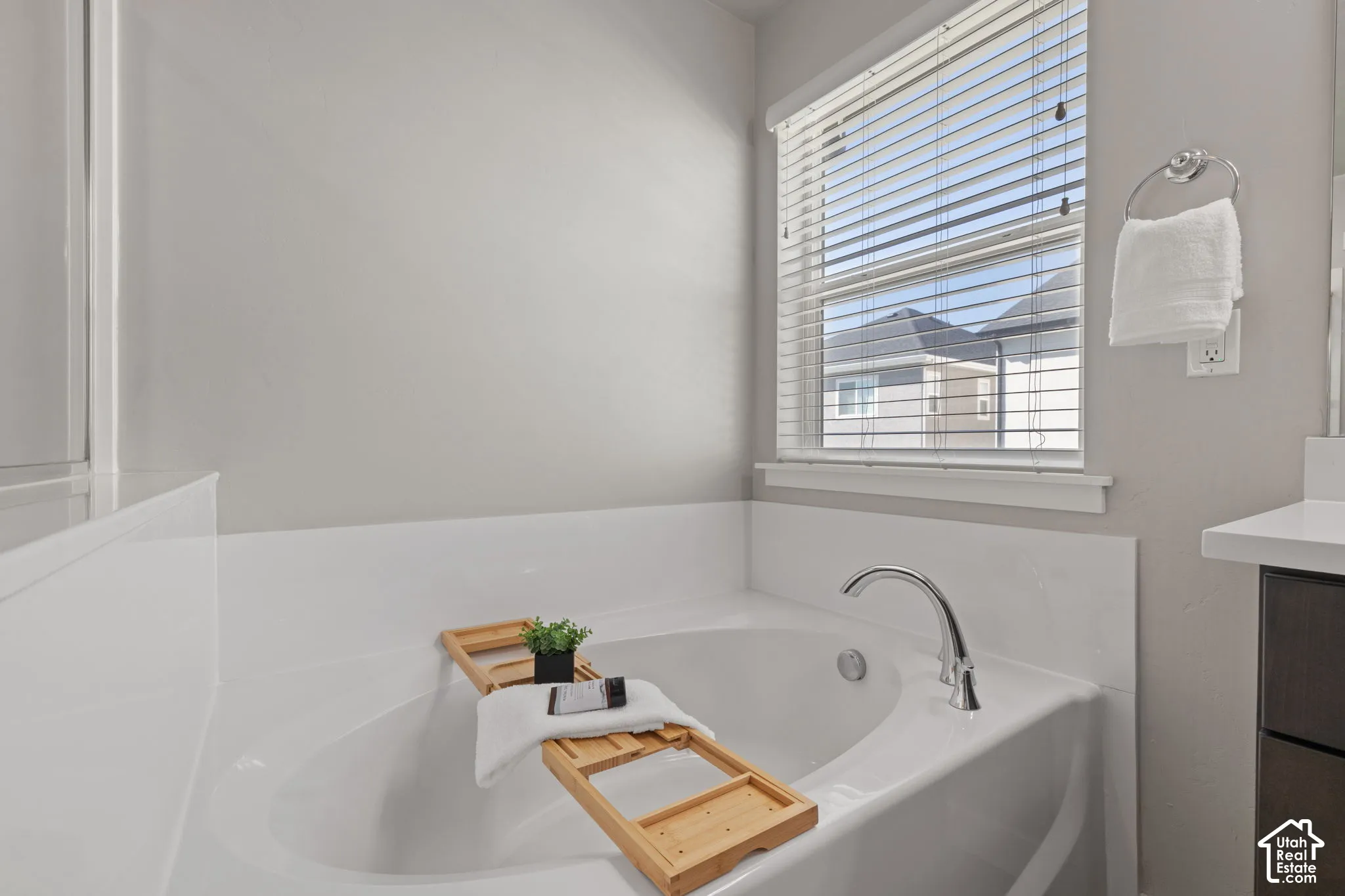 Spa-inspired primary bathroom featuring a freestanding soaking tub with chrome faucet, white subway tile wainscoting, soft neutral wall finishes, and natural light from a large window with horizontal blinds