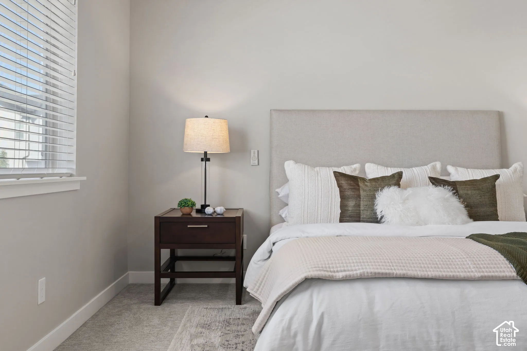 Upholstered headboard, neutral wall color, window with blinds, wood-finish nightstand, and carpeted flooring