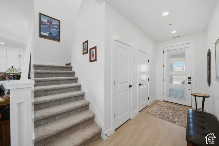 Light wood-finish flooring, two white panel doors with black hardware, a modern front door with frosted glass panels, recessed ceiling lighting, and a staircase with light gray carpeting