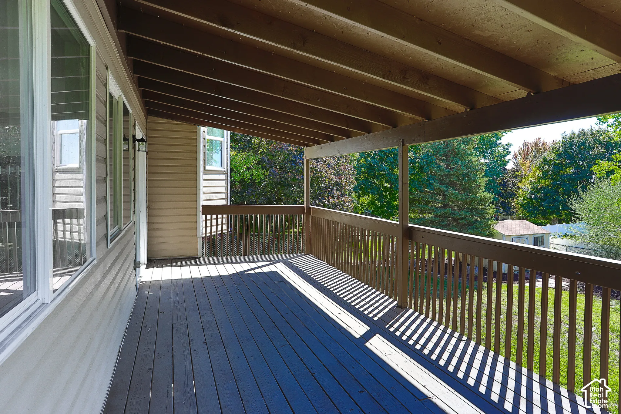 Expansive covered deck featuring a wood-finish floor and railing, exterior wall sconces, and multiple windows