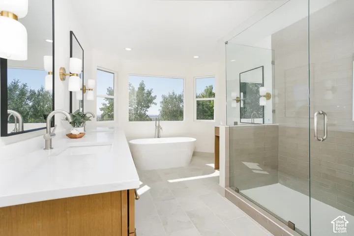 Dual sink vanity with white countertops, freestanding soaking tub, walk-in glass shower, and multiple windows