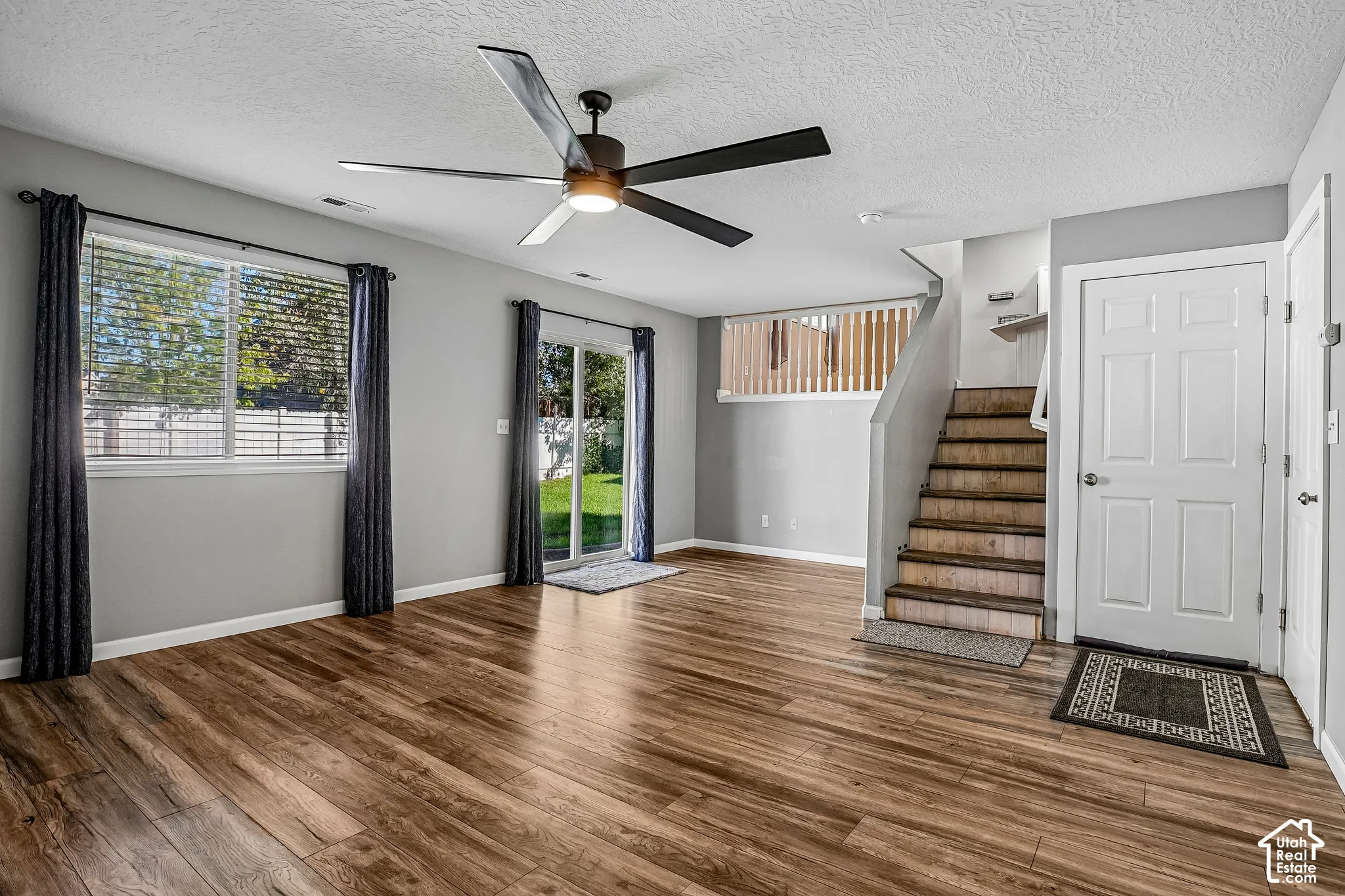 Wood-finish flooring throughout, ceiling fan with integrated lighting, glass sliding door to the exterior, multiple windows with blinds, and staircase with wood railing