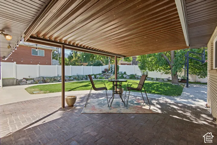 Covered patio with brick paver flooring, leading to a landscaped backyard featuring a white privacy fence and mature trees