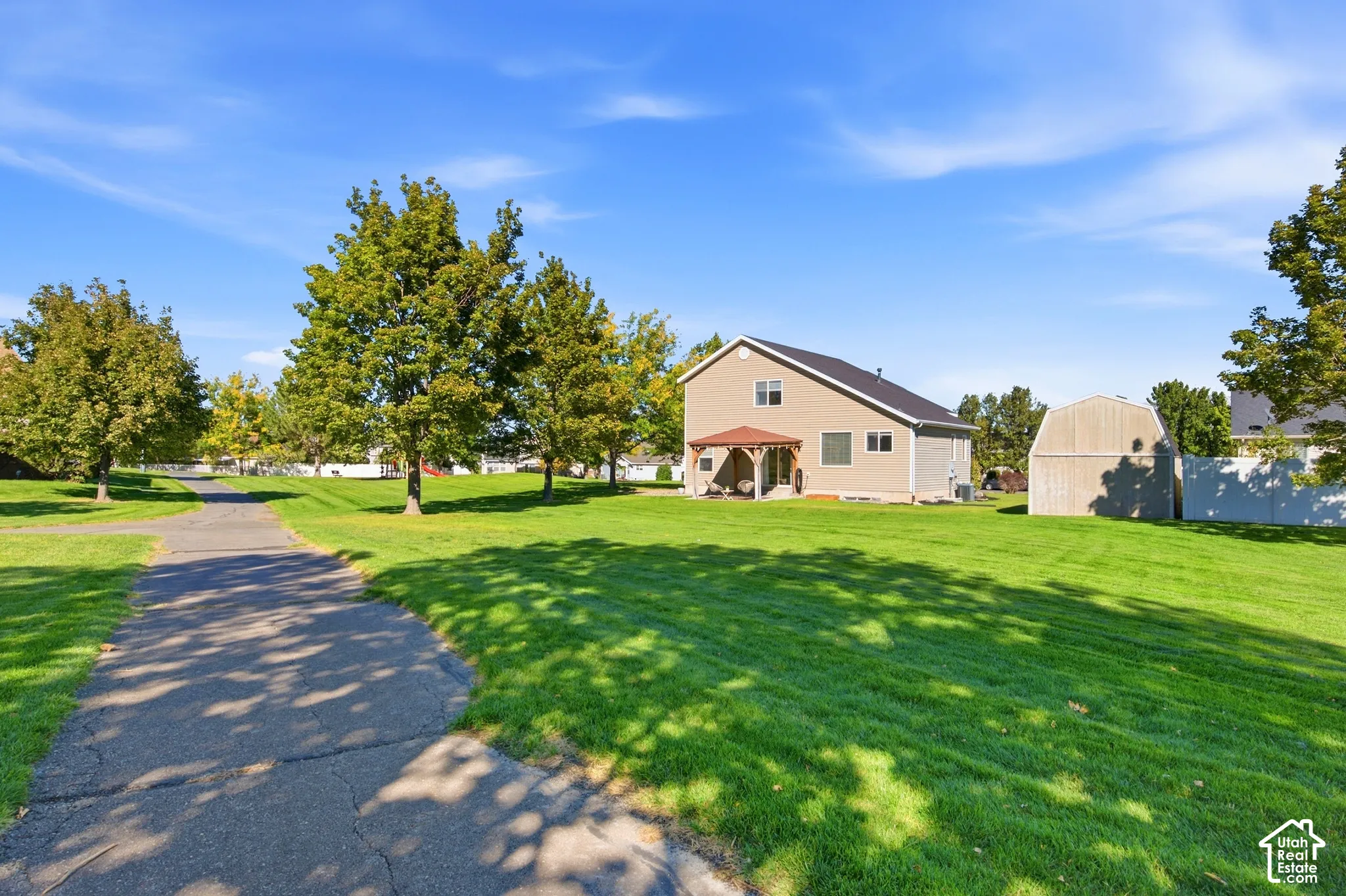 Expansive green lawn with mature trees, paved pathway, and a tan-sided residence featuring a covered patio