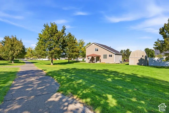 Expansive green lawn with mature trees, paved pathway, and a tan-sided residence featuring a covered patio