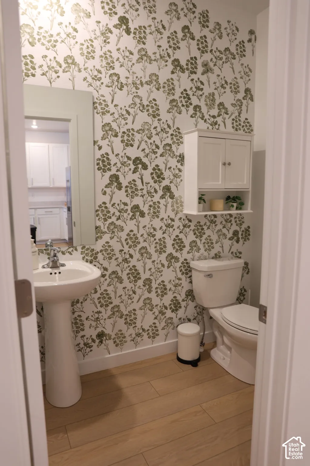 Pedestal sink with chrome faucet, decorative wall covering, wall-mounted storage cabinet, white toilet, and wood-finish flooring
