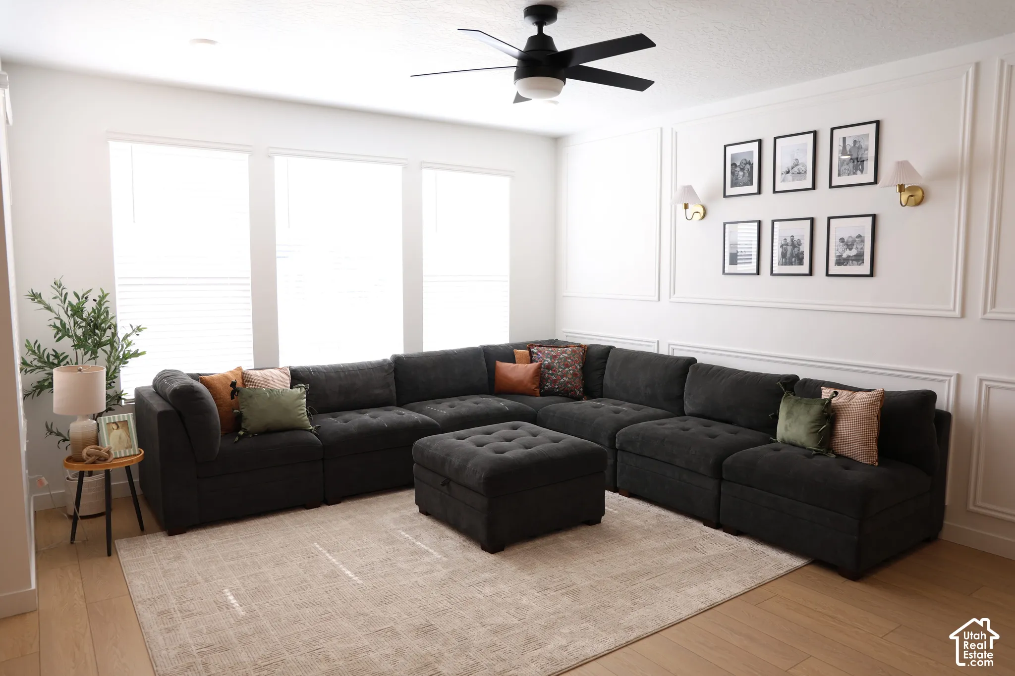 Wood-finish flooring, multiple large windows, decorative wall molding, black ceiling fan with integrated lighting, and wall-mounted sconces