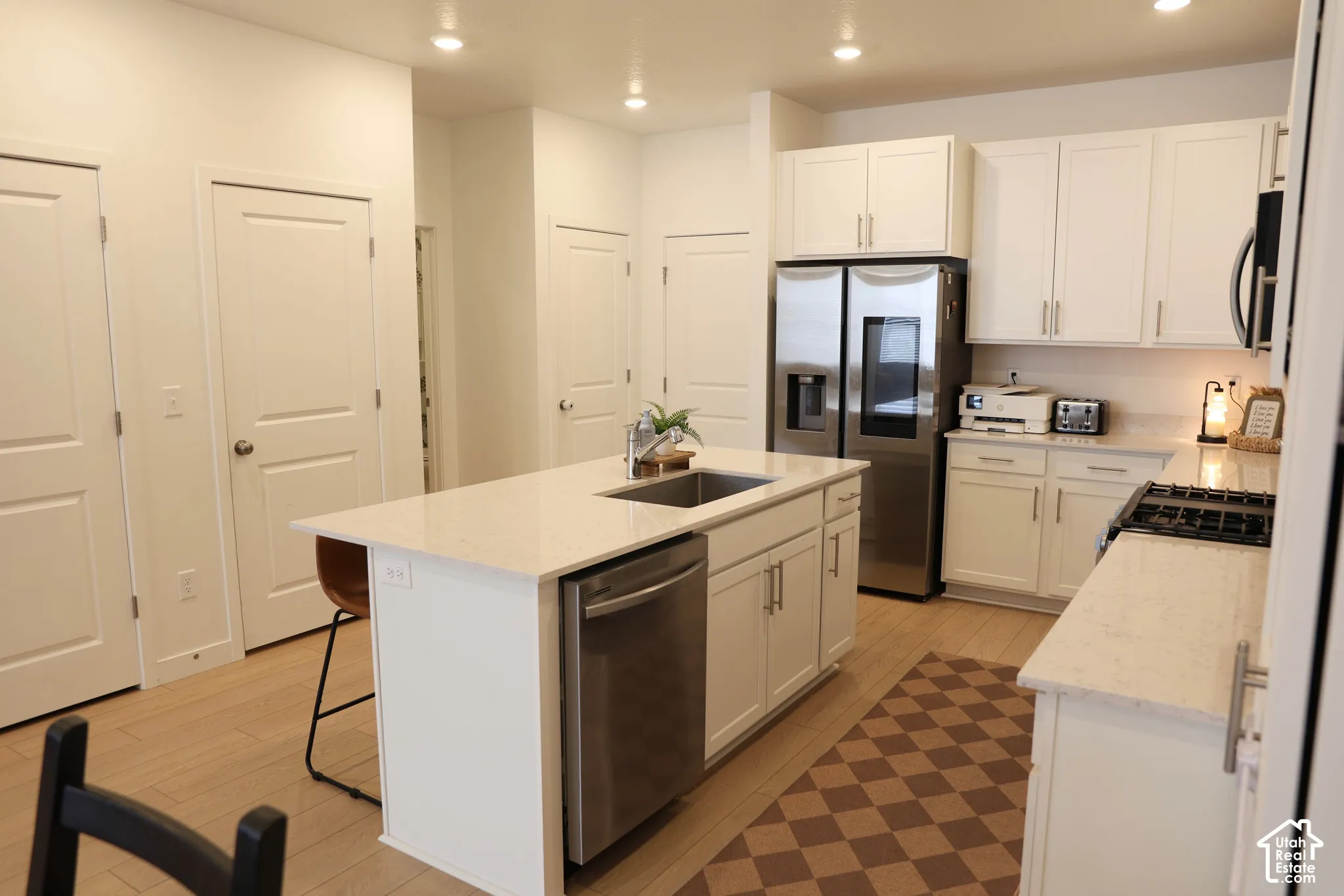 White shaker cabinetry, stainless steel appliances, central island with integrated sink, light-toned wood-finish flooring, and recessed lighting