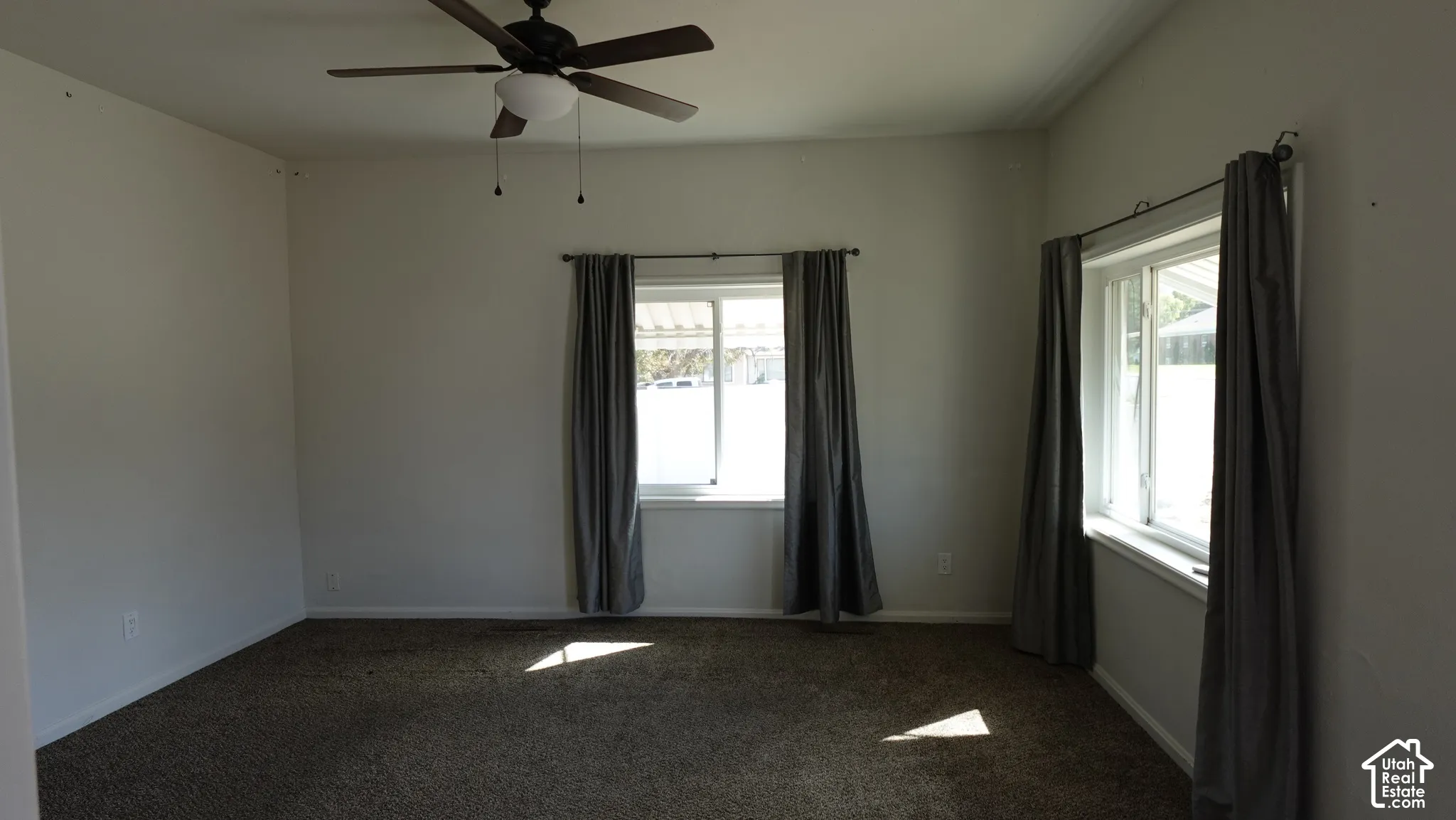 Bright, light-filled bedroom featuring dual windows with dark curtain panels, ceiling fan with pull-chain controls, and dark carpet flooring throughout