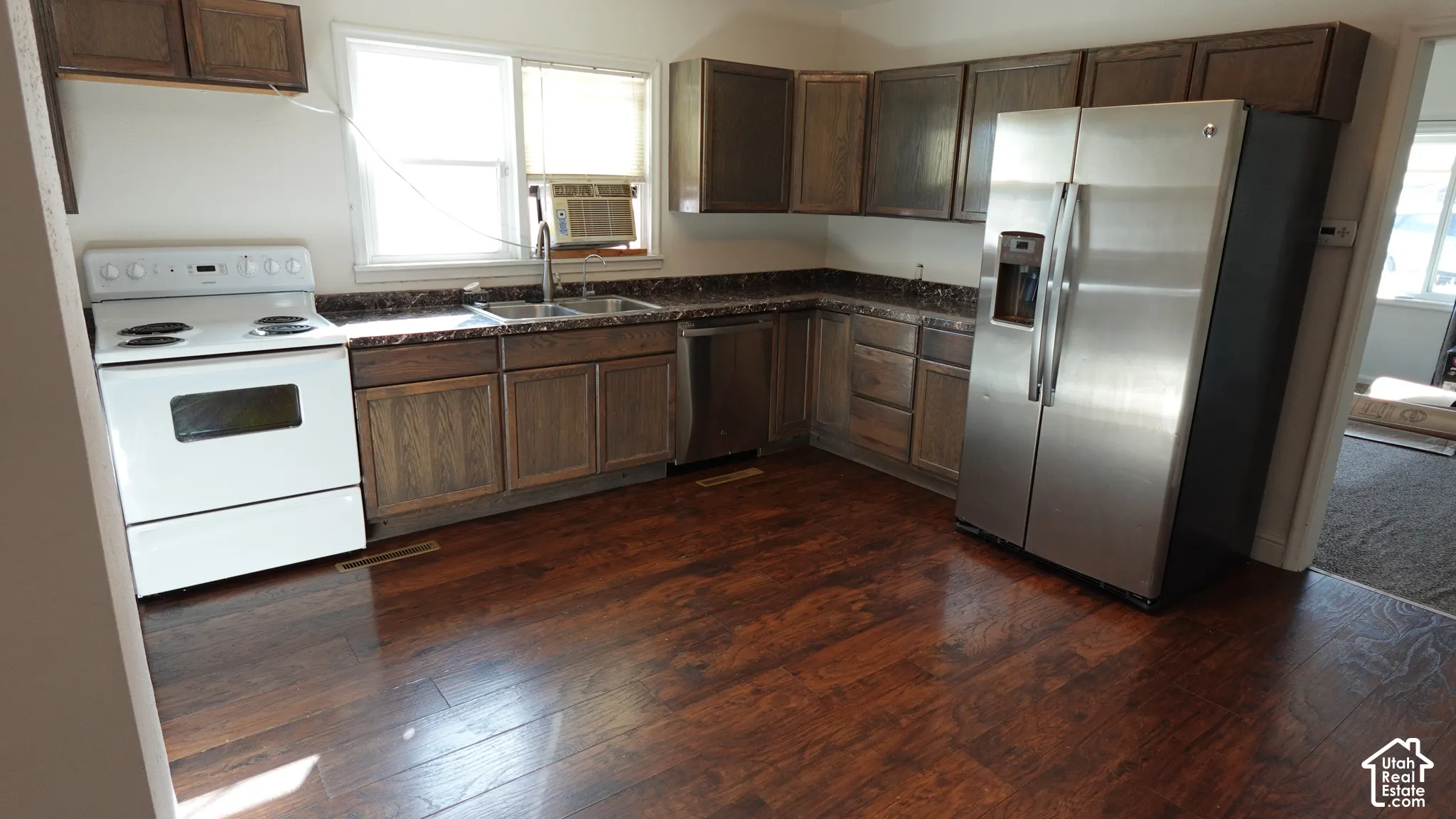 Spacious kitchen featuring wood cabinetry, stainless steel appliances including a side-by-side refrigerator, white electric range, and dishwasher, dark wood-finish flooring, and ample counter space with dark countertops throughout the L-shaped layout