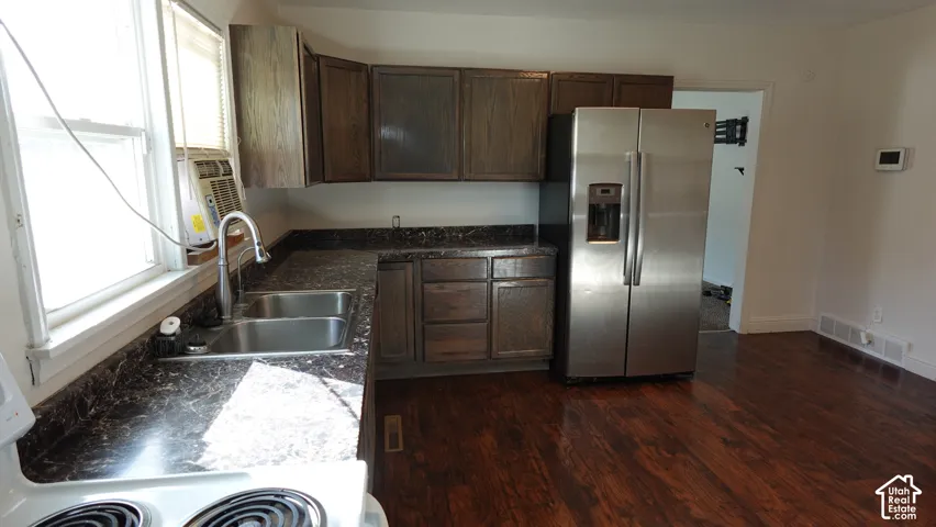 Chef's kitchen featuring dark wood cabinetry, stainless steel side-by-side refrigerator with ice and water dispensers, granite-style countertops, stainless steel sink with gooseneck faucet, and warm wood-finish flooring throughout the open layout