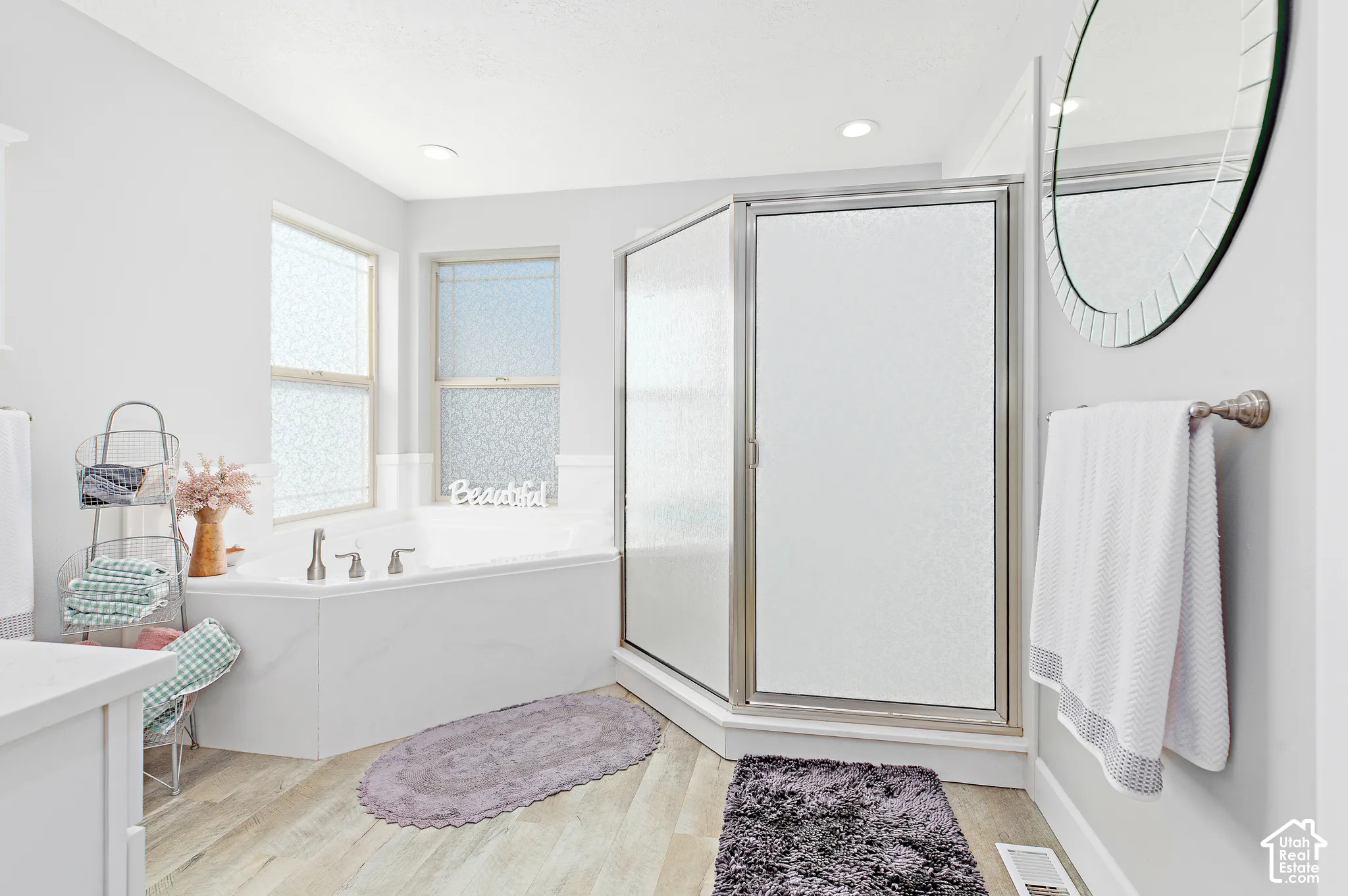 Corner soaking tub with dual windows, glass-enclosed shower, wood-finish flooring, and recessed ceiling lighting