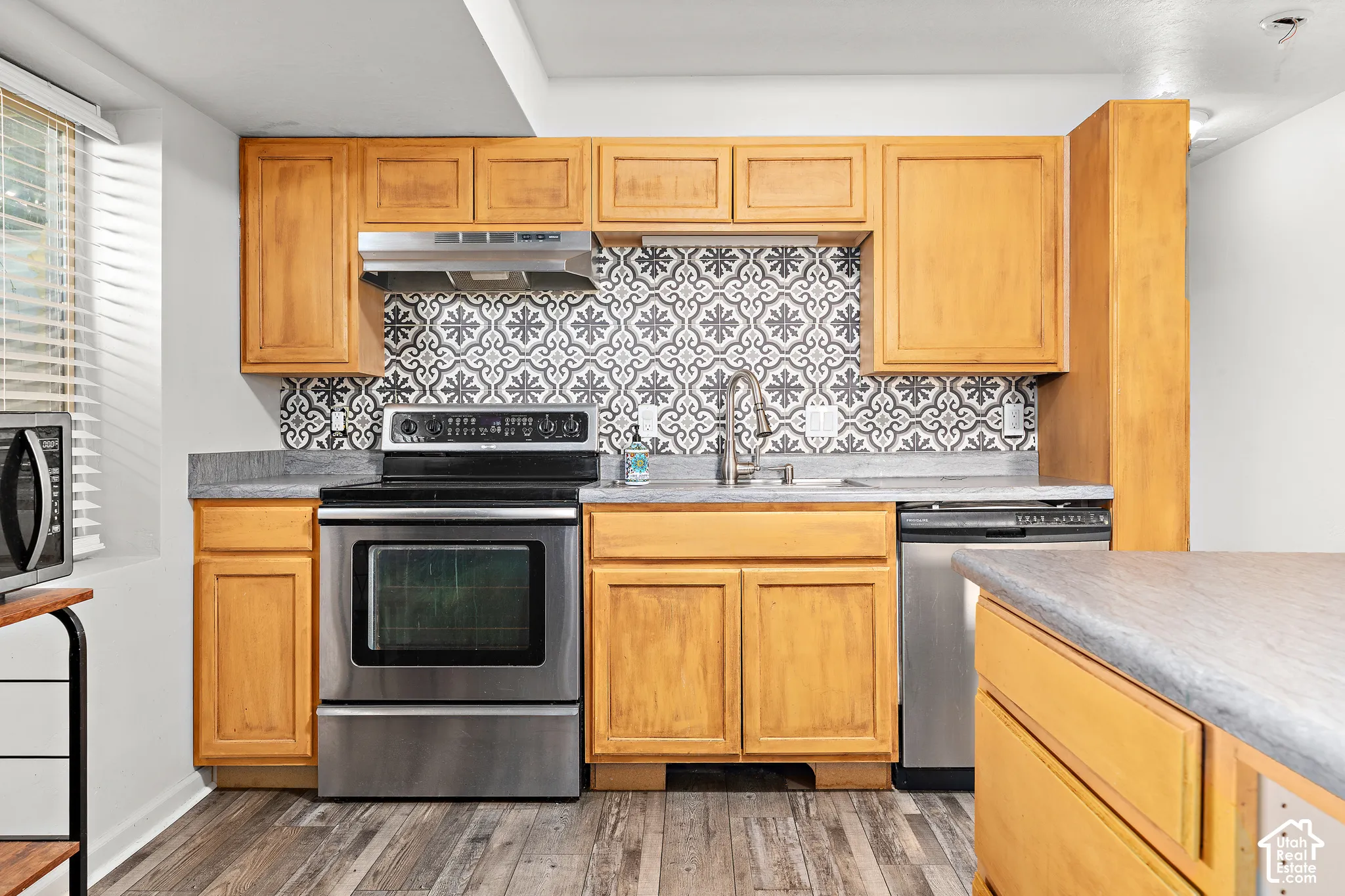 Wood-finish flooring with light wood cabinetry, a patterned tile backsplash, stainless steel appliances, and a double basin sink with a gooseneck faucet