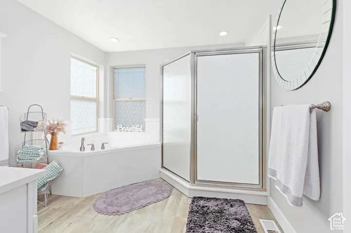 Corner soaking tub with dual windows, glass-enclosed shower, wood-finish flooring, and recessed ceiling lighting