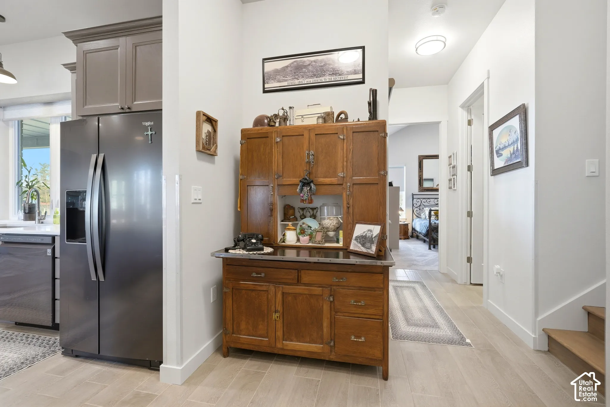 Stainless steel refrigerator, gray upper cabinetry, light wood-finish flooring, white walls, and a wood staircase