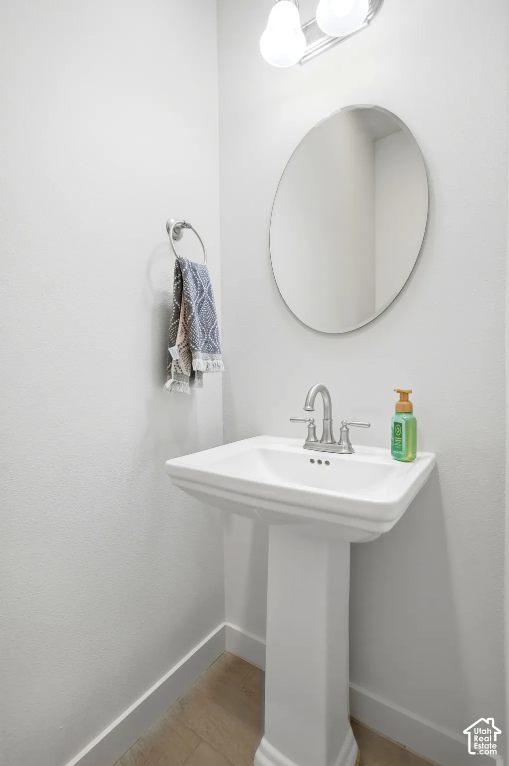 White pedestal sink with a chrome two-handle faucet, oval wall mirror, and dual-bulb vanity light