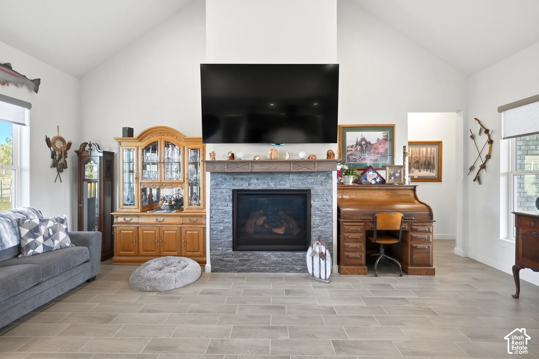 Vaulted ceilings, a stacked stone fireplace with a wood mantel, light-toned wood-finish flooring, and large windows with roller shades