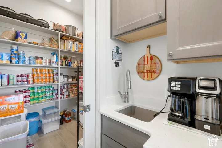 Expansive pantry with built-in shelving, coupled with a wet bar featuring a single-basin sink, gooseneck faucet, light grey cabinetry, and a light-toned countertop