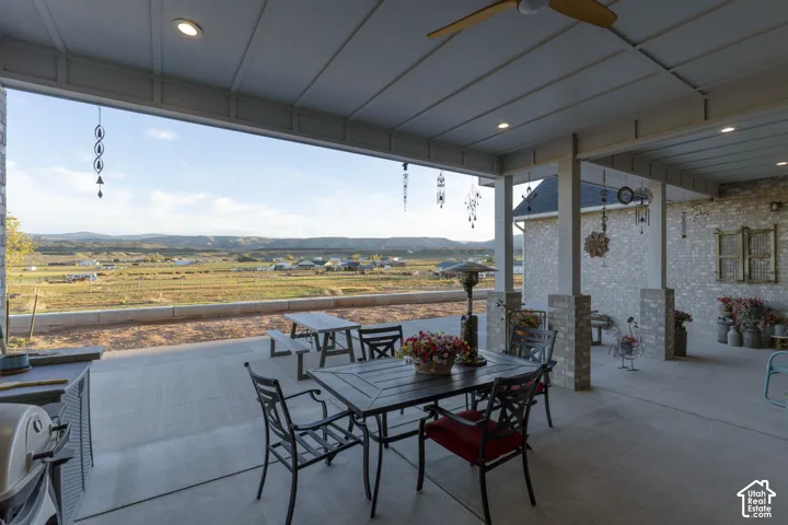 Spacious covered patio with a ceiling fan and recessed lighting, featuring a concrete surface and brick columns, overlooking distant terrain