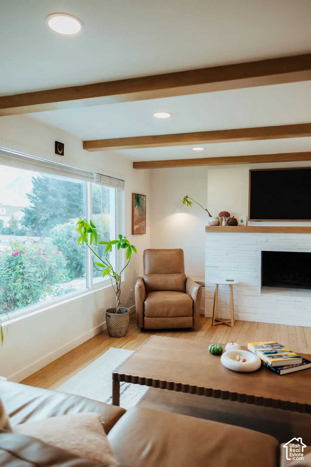 White-painted brick fireplace with a wood mantel, exposed wood ceiling beams, wood-finish flooring, and recessed lighting