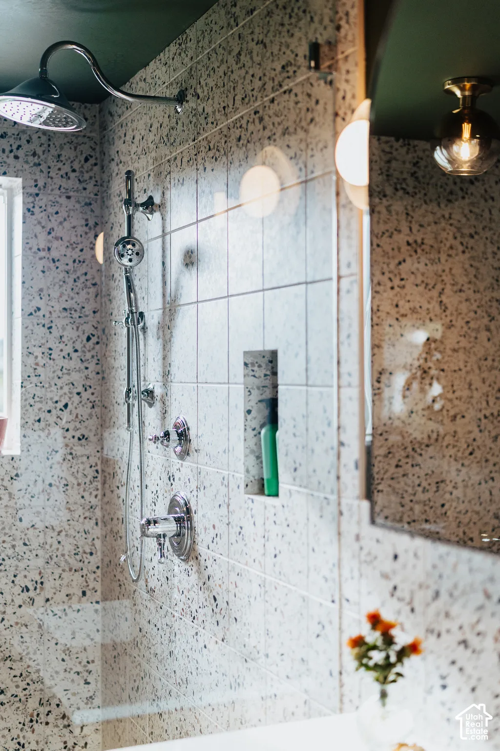 Shower enclosure featuring speckled tile surround, chrome multi-function shower system, built-in recessed storage niche, and a flush-mount brass globe light fixture