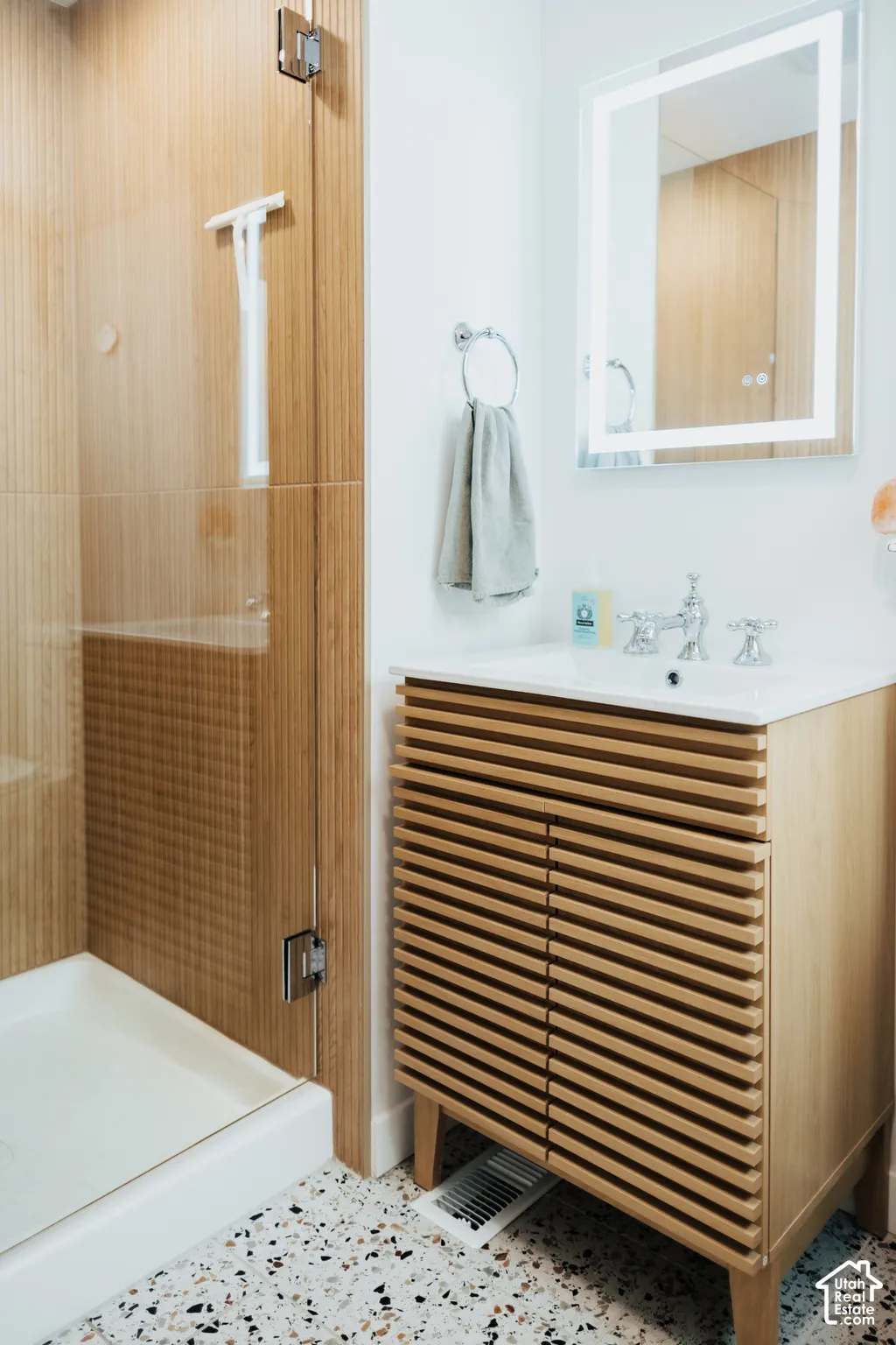 Slatted wood-finish vanity with an integrated countertop sink, LED-backlit mirror, glass-enclosed shower with vertical-tile surround, and speckled stone-finish flooring