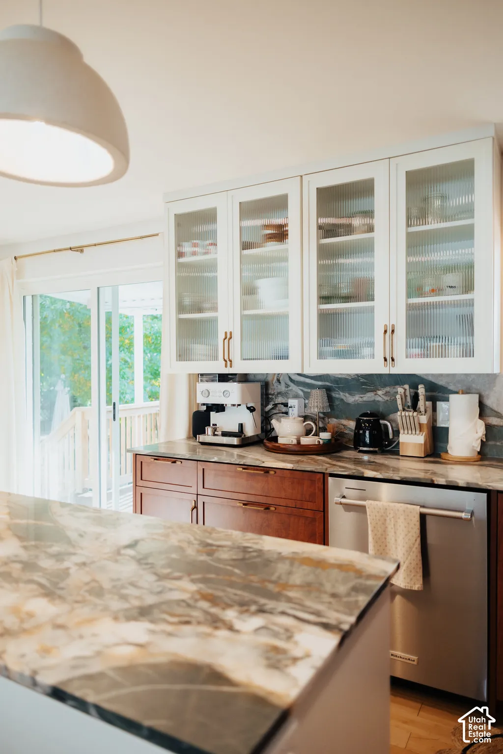 Kitchen featuring two-tone cabinetry with upper glass-front cupboards, natural stone countertops and backsplash, stainless steel dishwasher, and oversized dome pendant lighting