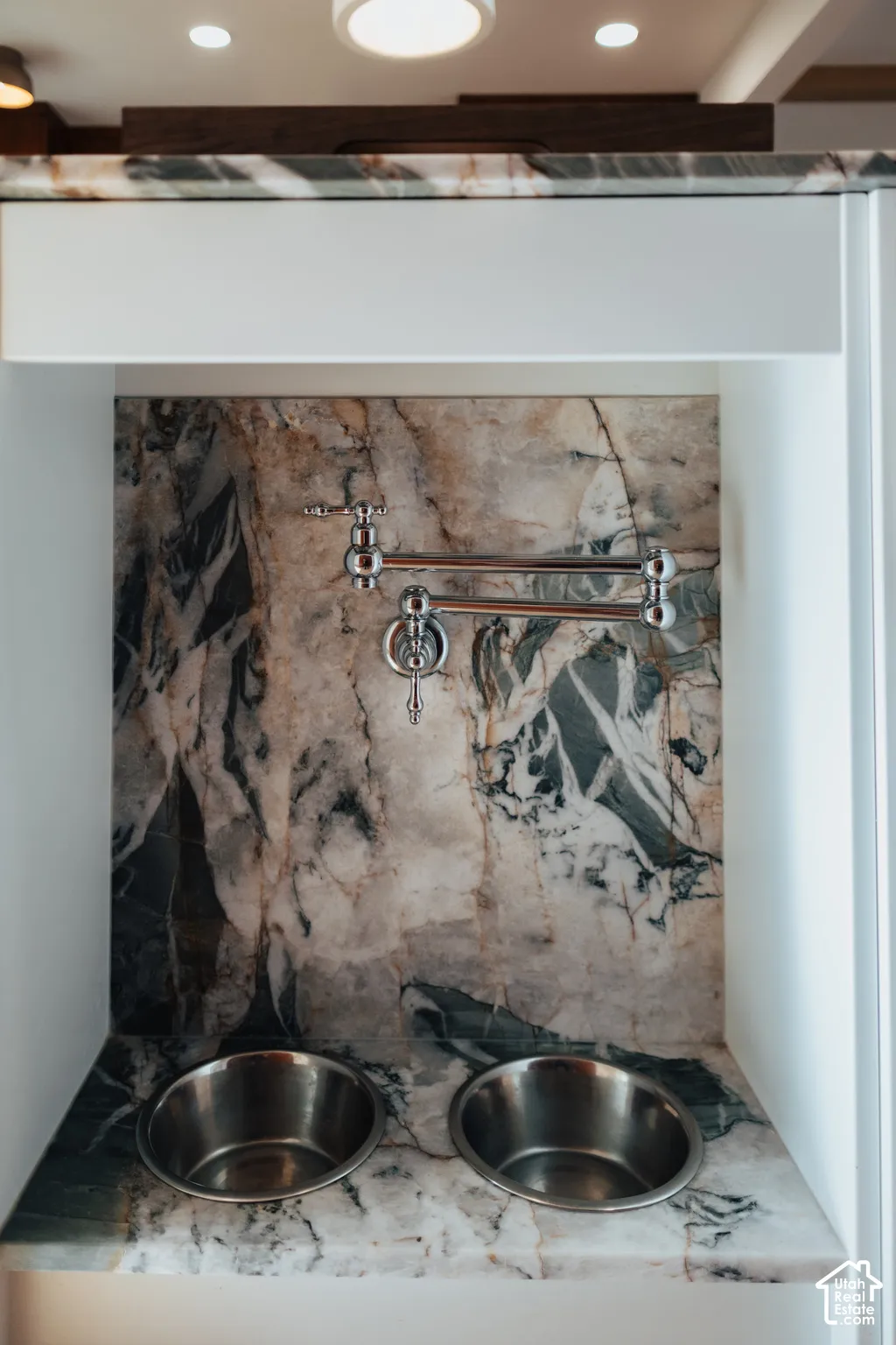 Custom alcove featuring a stone countertop and matching full-height backsplash, polished chrome articulated wall-mounted faucet, and two integrated stainless steel basins