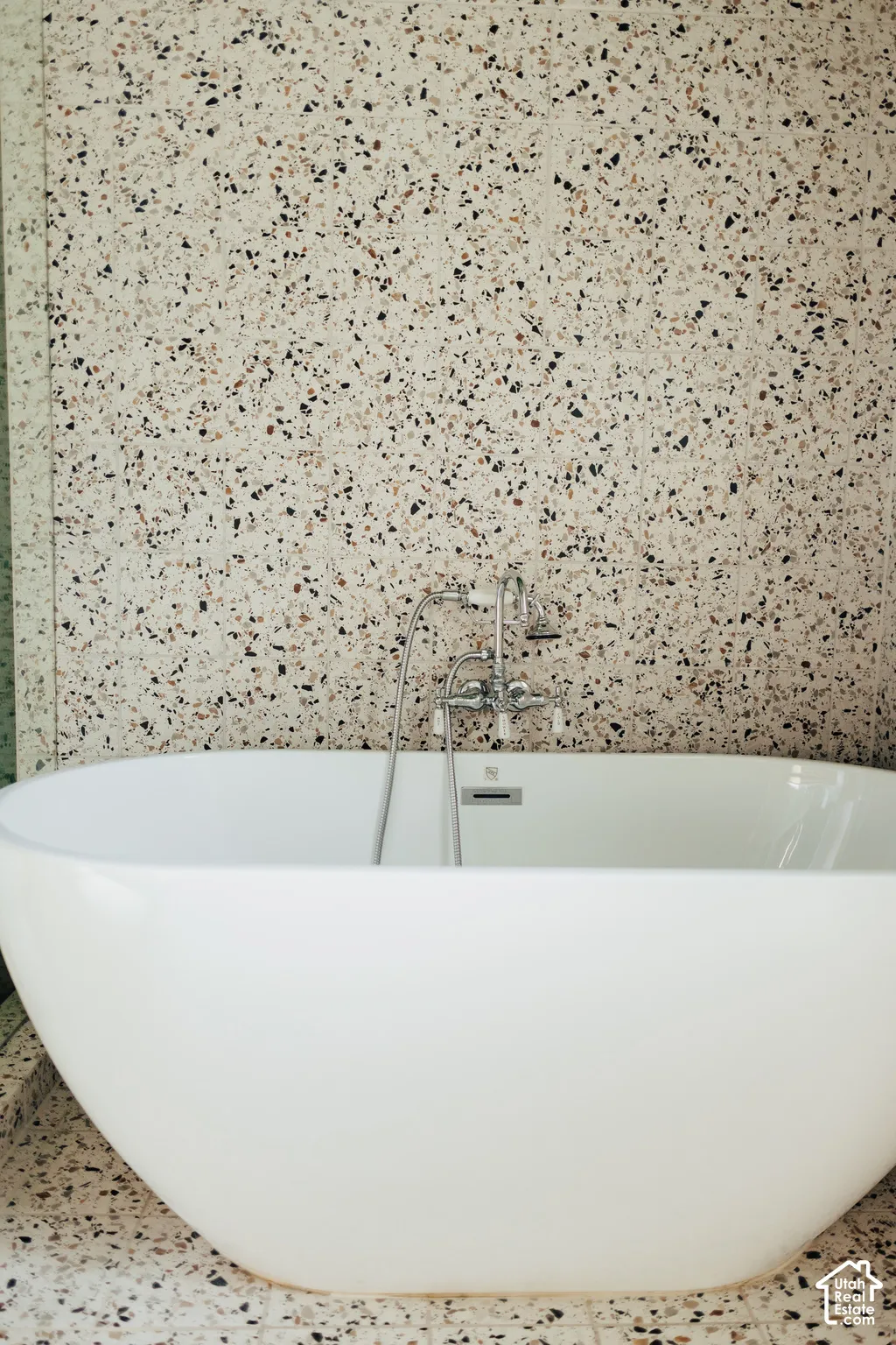 Freestanding white soaking tub with polished chrome deck-mounted faucet and handheld shower sprayer, set against full-height terrazzo wall and floor tiling
