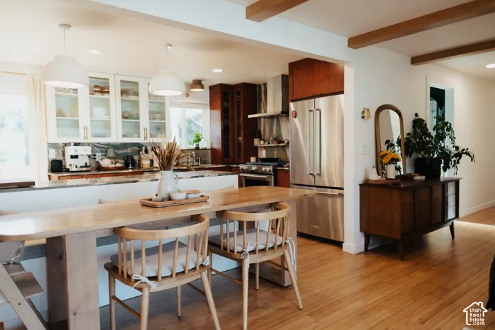 Kitchen featuring two-tone cabinetry with glass-front uppers, stainless steel refrigerator, stone-surfaced island, wood-finish flooring, and exposed ceiling beams